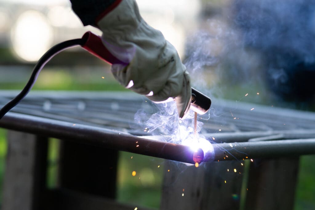 A close-up of a mobile welder expertly welding metal bars, with sparks and smoke dancing in the air. Wearing protective gloves and wielding a torch, the skilled artisan operates against a blurred backdrop of greenery, evoking a sense of plant repair in motion.