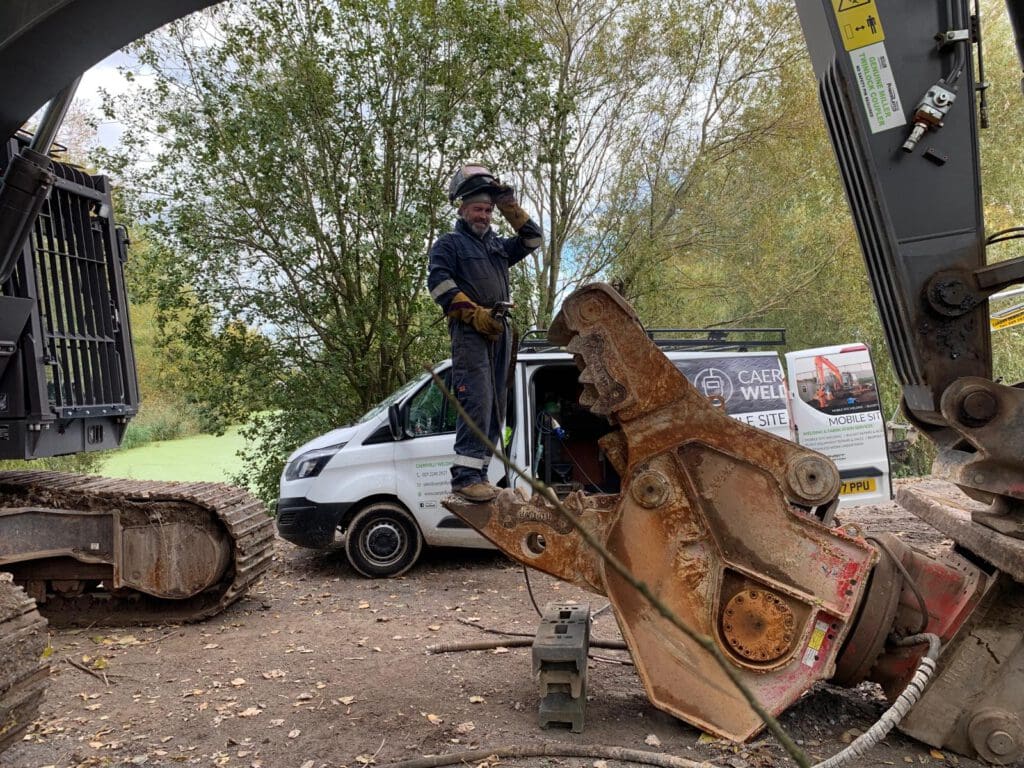 A man wearing protective gear stands on an excavator attachment, appearing to adjust his helmet. A white van with signage and trees are in the background, suggesting a construction site.