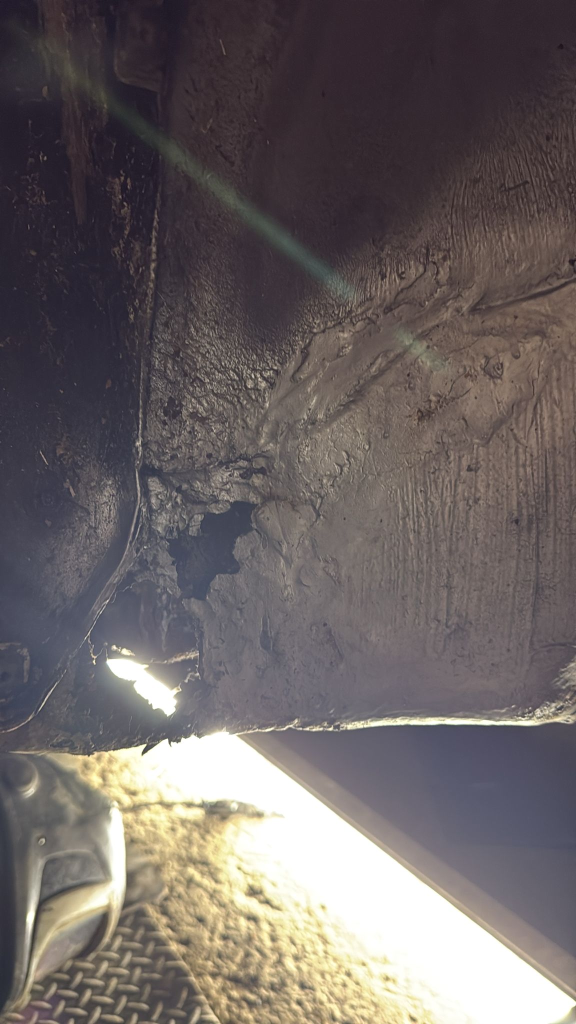 Close-up view of a damaged, rusted section of a metal surface, possibly part of a vehicle undercarriage, with a bright light source illuminating the area and highlighting cracks and rough textures.